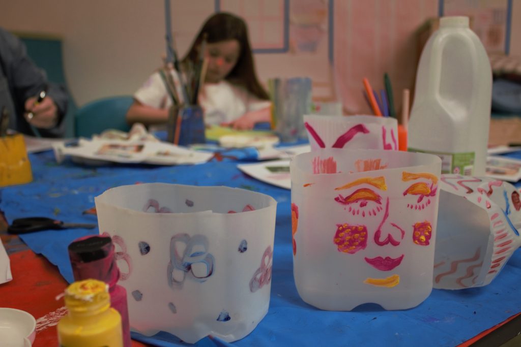 Painted recycled milk carton flower pots sit in the foreground. In the background a girl can be seen painting her pot.